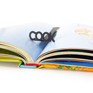 Knife-shaped COOK metal bookmark standing upright in the center of an open illustrated book with colorful pages and blue background.