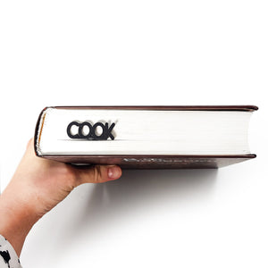 Closed book held by hand with the COOK knife-shaped metal bookmark visibly sticking out from the top edge.