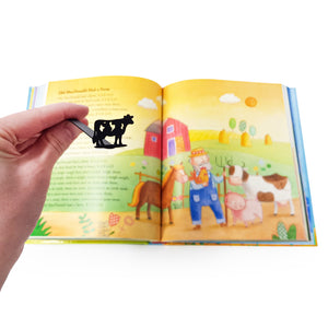 Hand holding metal cow bookmark over open children's book showing Old MacDonald farm scene