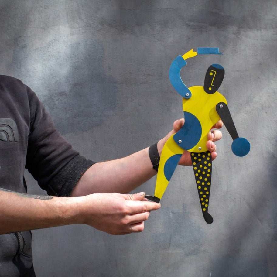 Craftsman holding yellow and blue Bauhaus jumping jack puppet against grey wall
Image