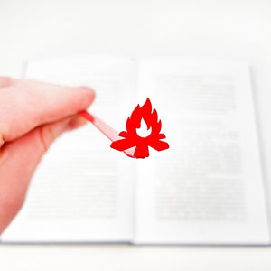 Red bonfire metal bookmark held in hand above open book, minimalist steel page marker with smooth powder coated finish