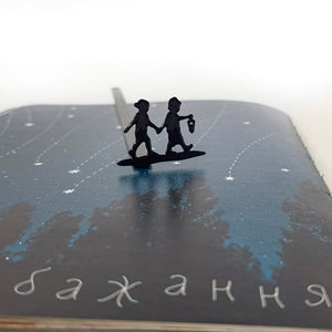 Black metal bookmark with two children holding hands, one carrying a lantern, placed inside a book opened to an illustrated night sky with stars and Ukrainian text.