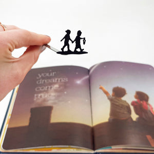 A hand holding a black metal bookmark featuring two children walking hand in hand with a lantern, in front of an open book showing a dreamy scene of children stargazing and the words “your dreams come true.”