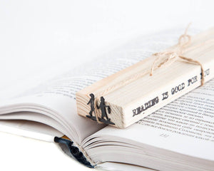 Black metal bookmark shaped like two children holding hands, attached to a wooden block stamped with the phrase “Reading is good for you,” resting on an open book.