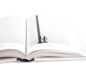 Black metal bookmark placed in the center of an open book, featuring a silhouette of two children walking hand in hand, one holding a lantern.