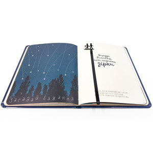 Black metal bookmark with two children holding hands and a lantern, placed inside an illustrated journal showing a starry night sky and Ukrainian text about making wishes.