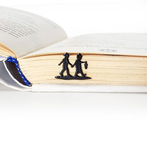 Close-up of a black metal bookmark shaped like two children holding hands, one carrying a lantern, placed between the pages of a book so only the silhouette is visible at the book’s edge.