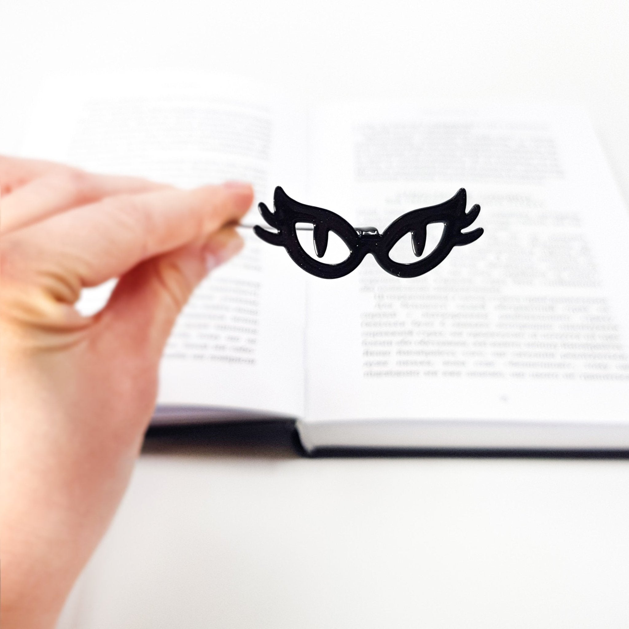 Black metal cat’s-eye glasses bookmark held above an open book, playful masquerade-style design for book lovers. The bookmark is handcrafted in Cherkasy, Ukraine. It is not mass-produced.