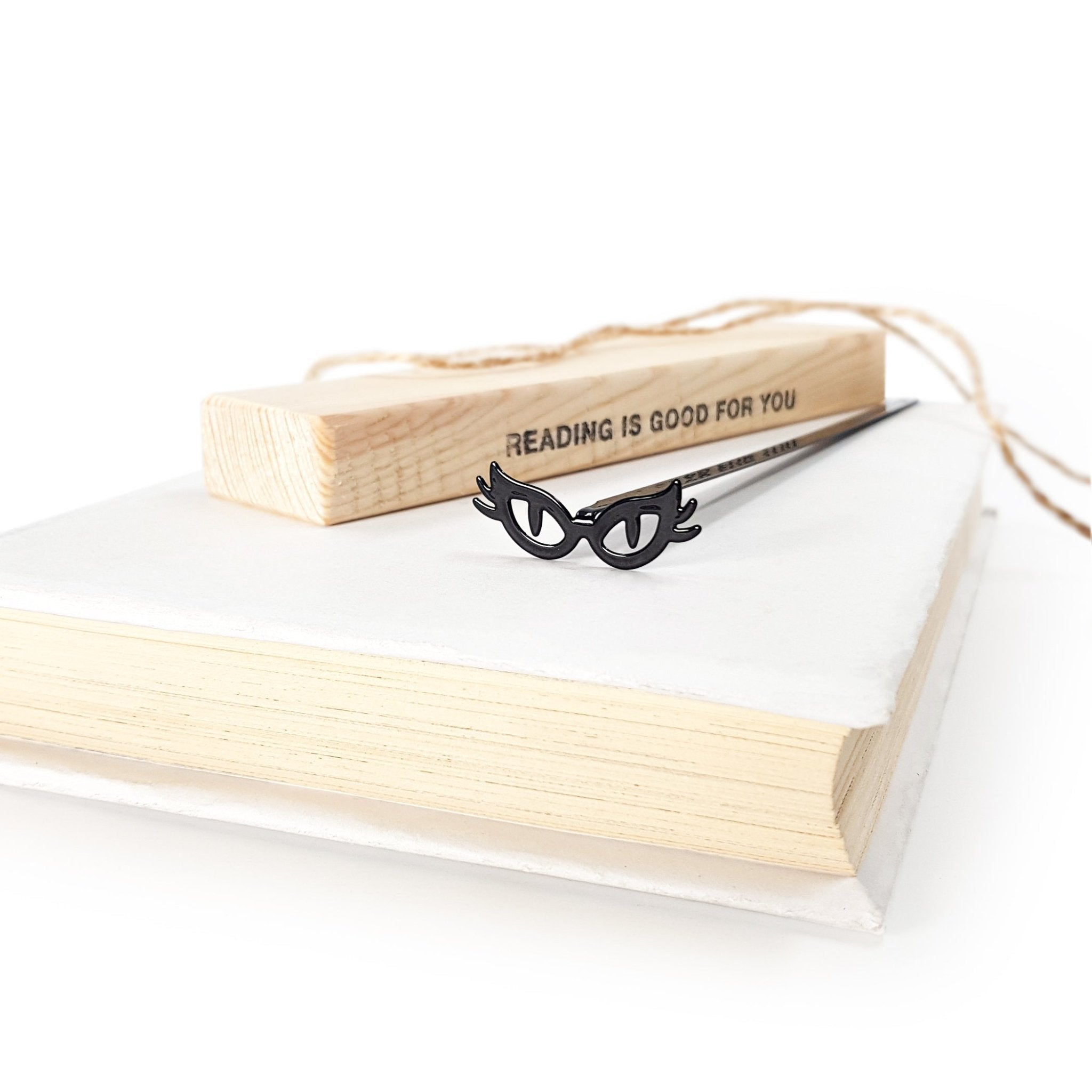 Black metal bookmark shaped like cat’s eye glasses resting on a closed book, with a wooden block reading “Reading is good for you” in the background.