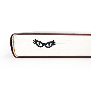 Black metal cat-eye glasses bookmark resting against the page edges of a closed book, creating a subtle peeking eyes effect for book lovers.