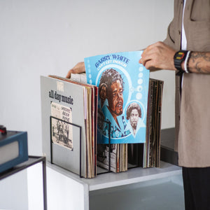 Person browsing vinyl records stored in a minimalist black metal holder; prominently featured is the colorful “Barry White – Can’t Get Enough” album cover, set on a light grey shelf.