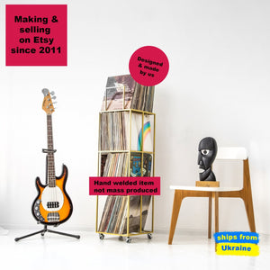 Three-quarter view of a gold metal Four - tier Vinyl Record Stand on Rubber Wheels with records near a guitar and a black head statue reminiscent of a cover of Division Bell by Pink Floyd.