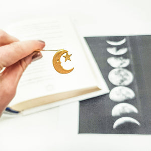 Hand holding a gold crescent moon and star bookmark in front of an open book and a black and white moon phase chart