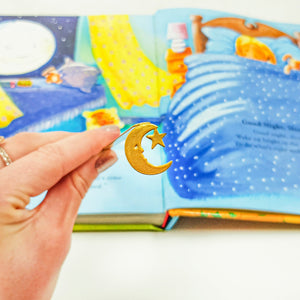 Hand holding a gold crescent moon and star bookmark in front of an open illustrated bedtime storybook with a cozy bedroom scene