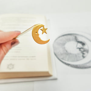 Gold crescent moon bookmark with star cutout, held above an open book and vintage moon illustration in soft background. Designed and made by Atelier Article in Ukraine. Not mass-produced.