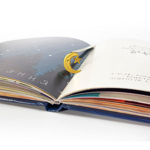 Gold crescent moon and star bookmark clipped to the center of an open illustrated book with Ukrainian text and celestial imagery