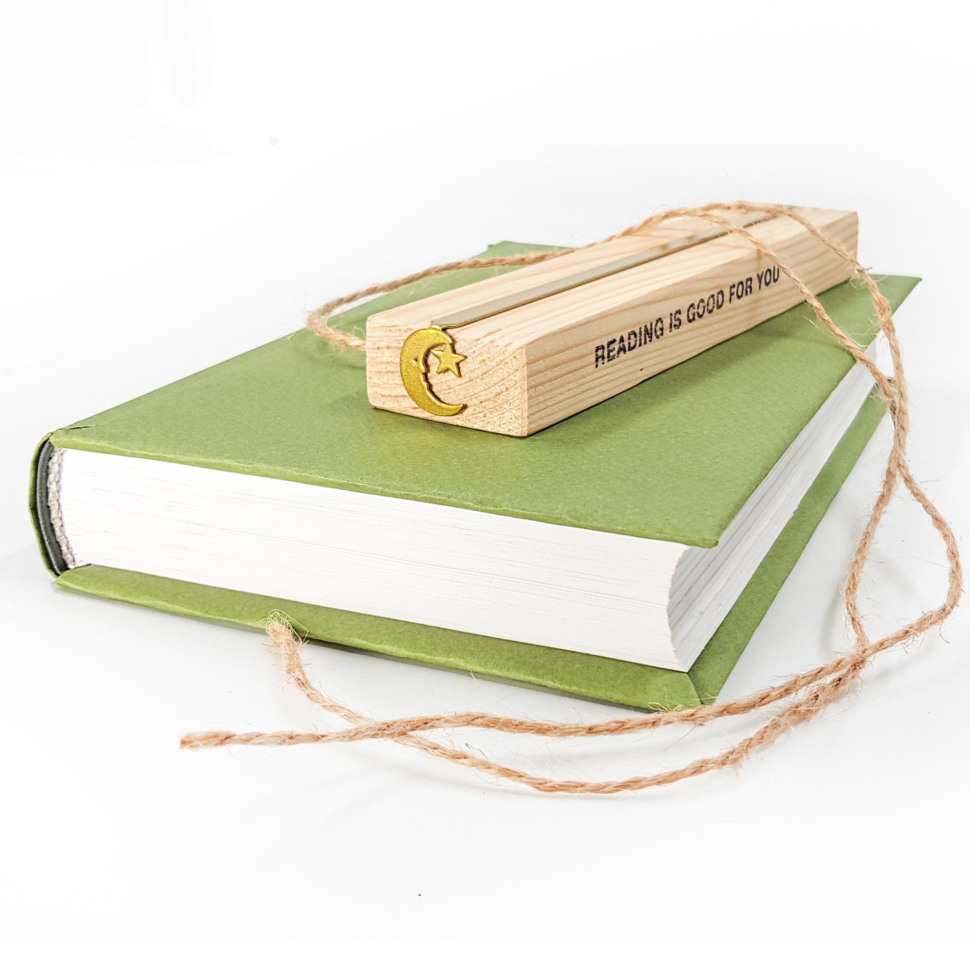 Gold moon and star bookmark packed in a wooden block engraved with ‘READING IS GOOD FOR YOU’, resting on a green hardcover book with twine.