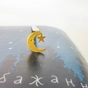Gold crescent moon and star bookmark clipped to a book page with a starry night sky illustration and Cyrillic text at the bottom