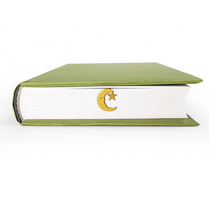 Gold crescent moon and star bookmark peeking out from the closed pages of a green hardcover book lying flat on a white background