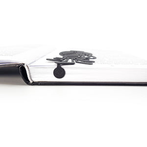 Side view of a black metal golf bag bookmark resting between book pages, showing the thin profile and small rounded tab extending below the page edge.