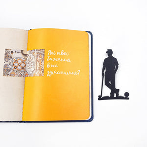 Black metal golf player bookmark placed next to an open notebook with yellow pages, showing size and silhouette design.