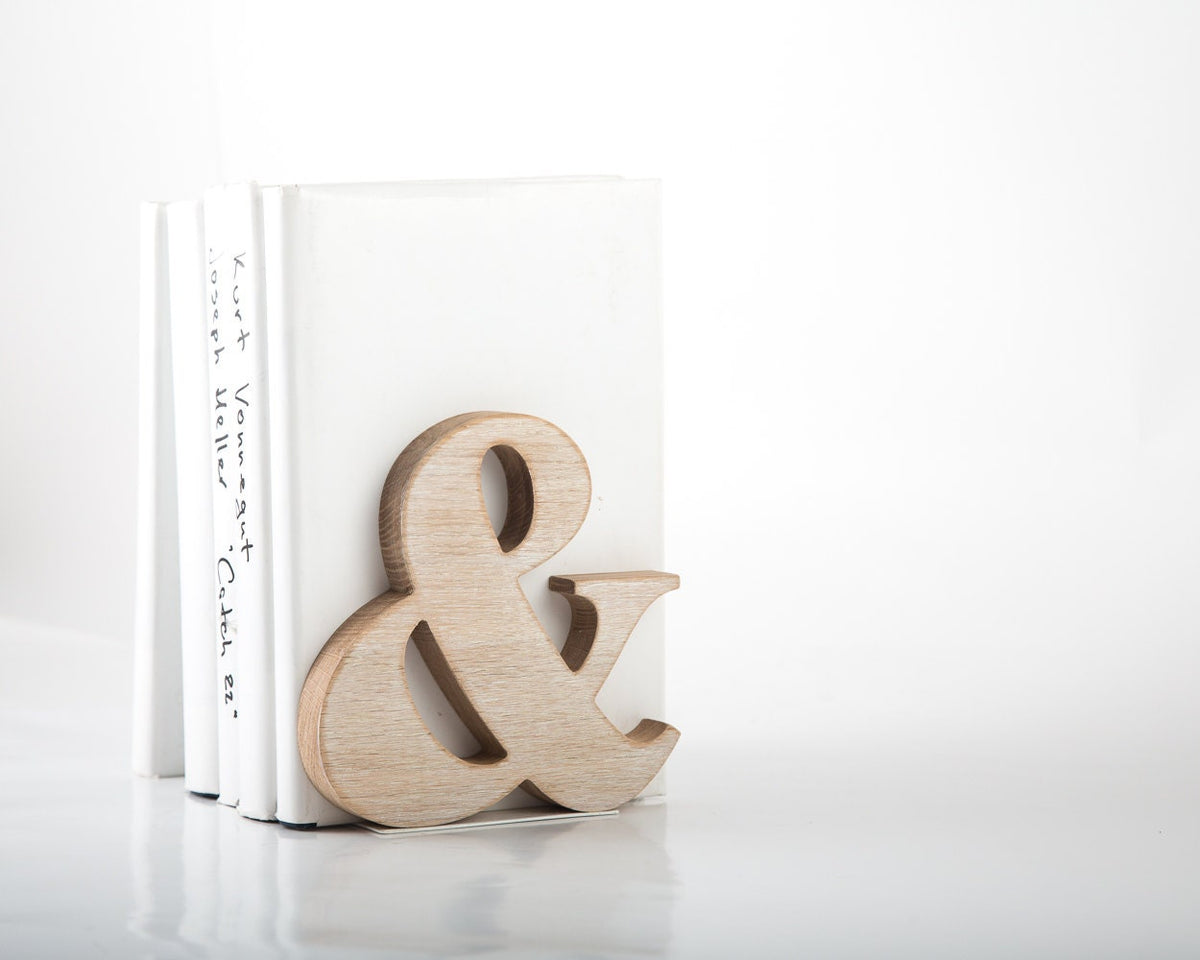 Natural wood ampersand symbol standing on white surface with stacked books in background, minimalist decorative accent for modern shelf or desk.
