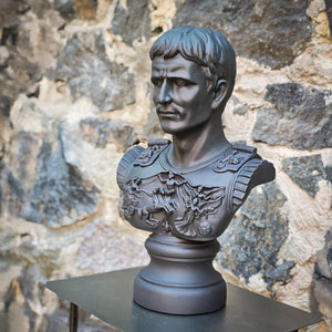 A dark gray plaster bust sculpture of a Roman emperor Julius Caesar with military armor and a lion emblem, displayed on a black table against a rustic stone wall background.