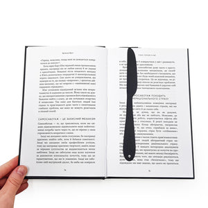 Black metal knife-shaped COOK bookmark placed vertically in an open book with Ukrainian text, held by a hand.