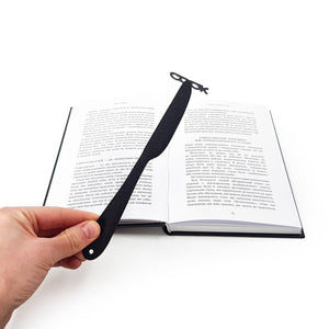 Hand holding a black knife-shaped COOK metal bookmark above an open book with Ukrainian text, pages spread flat on a white surface.