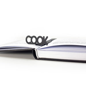 Knife-shaped COOK metal bookmark standing upright in the spine of an open book with black text, against a clean white background.
