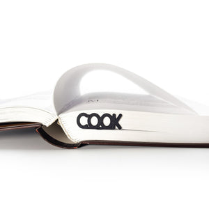 Black COOK metal bookmark inserted into the top of a partially opened book, with pages curled upward and white background.