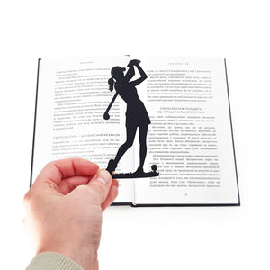 Hand holding a black metal lady golf player bookmark over an open book, showing the slim silhouette design marking the page