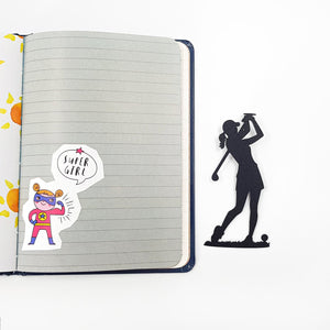 Black metal lady golf player bookmark placed next to an open notebook on a white background, showing the slim silhouette design