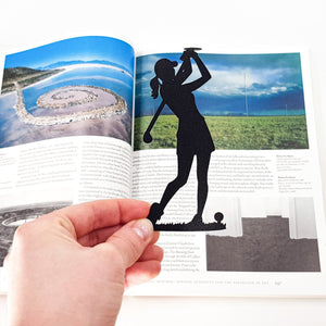 Hand holding a black metal lady golf player bookmark in front of an open art book, highlighting the golfer silhouette against the page