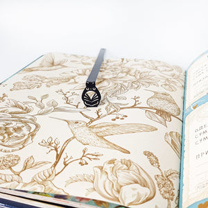 Black laser cut metal owl bookmark placed inside an open book with vintage botanical and bird illustrations, showing the slim matte black design.