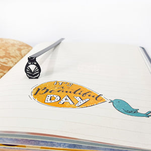 Black metal owl bookmark placed on an open notebook page with “It’s a Beautiful Day” illustration, showing the slim 6.8 inch laser cut design.