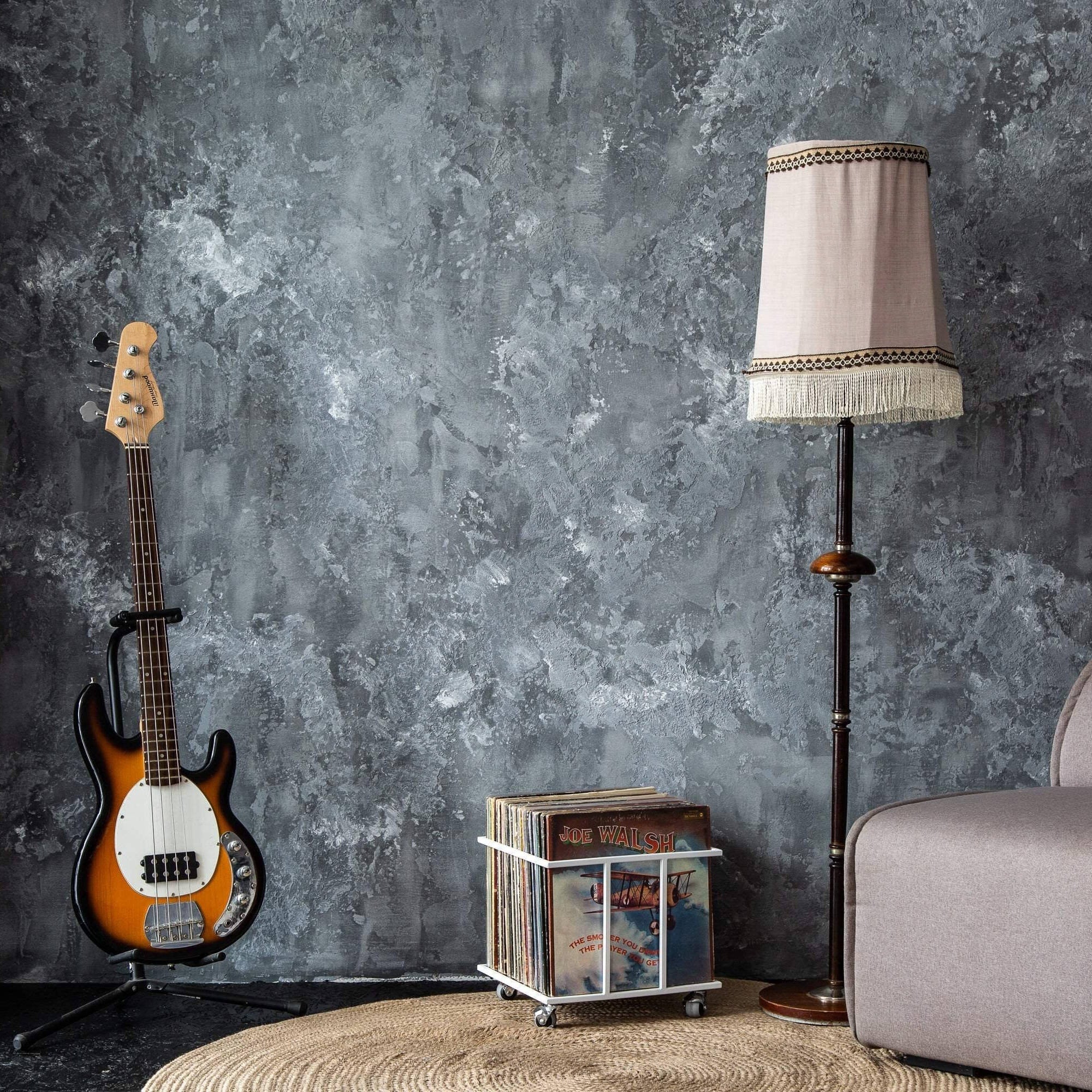 White metal vinyl record crate on wheels in a cozy music corner with a bass guitar, vintage lamp, and gray textured wall. Light Metal Crate on Wheel for Vinyl Records. Designed and made by Atelier Article