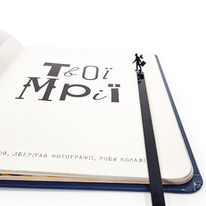 Black metal bookmark with a businesswoman silhouette standing at the top, placed inside an open notebook with minimalist typography.