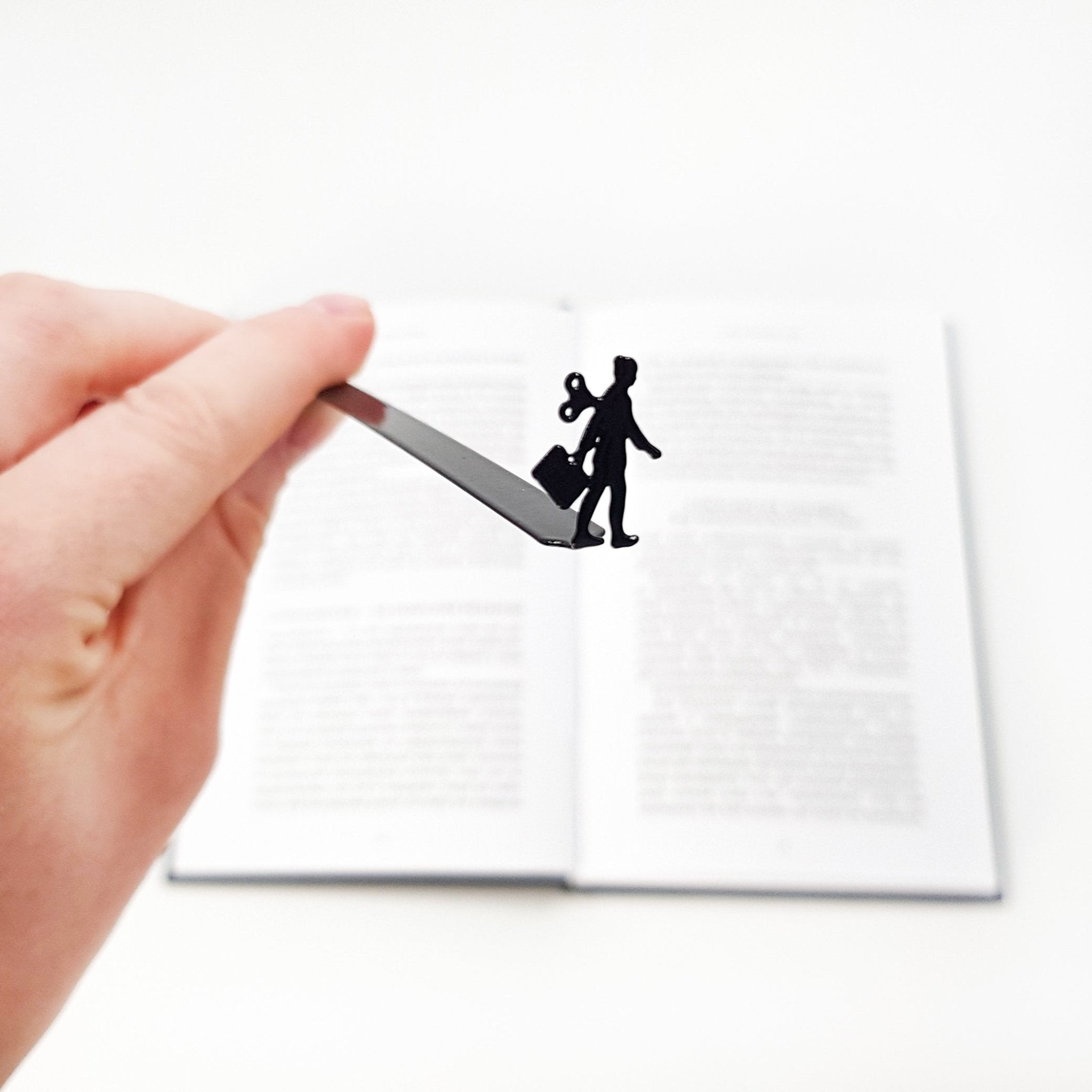 Hand holding a black metal bookmark shaped like a walking businesswoman with a wind-up key above an open book. The bookmark is hand-crafted in Cherkasy, Ukraine. It is not mass-produced.