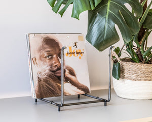 Minimalist steel vinyl record rack with tall open frame, holding LPs including “Joy” by Isaac Hayes, placed on a light surface next to a potted monstera plant.