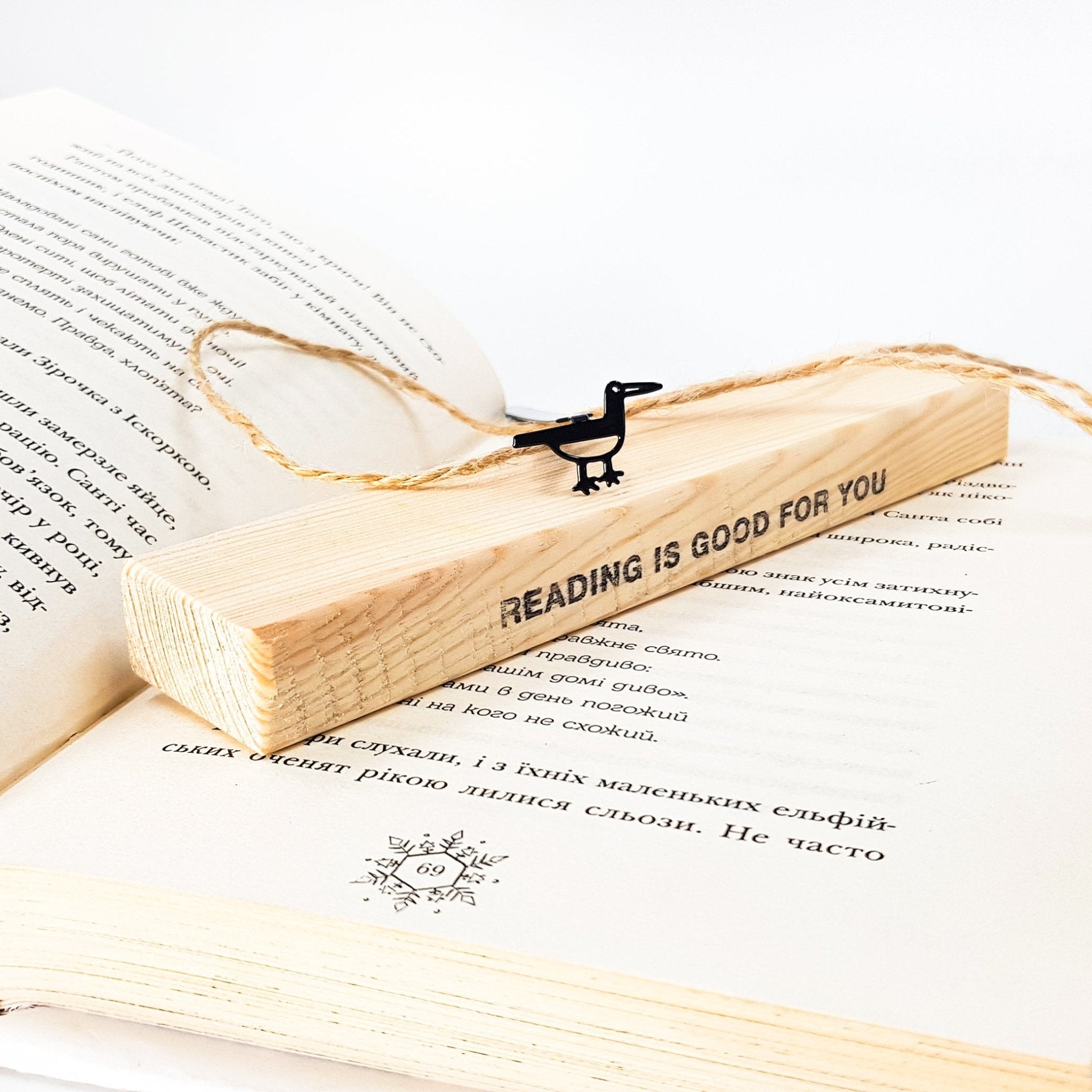 Minimalist black metal bird bookmark resting on a wooden block engraved “READING IS GOOD FOR YOU,” placed on an open book.