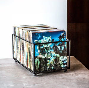 Metal vinyl record holder on wheels filled with colorful LPs, placed on a smooth concrete surface against a white and dark wood wall.