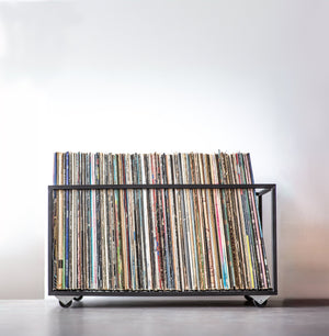 Low-profile black metal record storage bin filled with a large collection of vinyl records, neatly organized and displayed on a smooth surface against a white background.