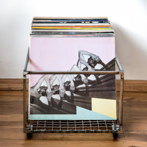 Metal vinyl record holder with wheels, filled with records, fronted by a large LP with abstract art of repeated human figures, placed on a wooden floor.