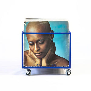 Blue metal vinyl record holder on wheels, showcasing a bold album cover with a close-up portrait of a bald woman against a turquoise background.