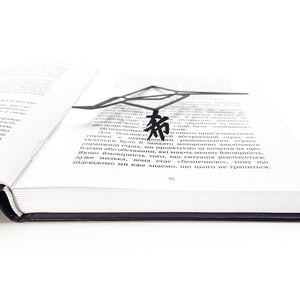Low-angle close-up of a black metal origami crane bookmark lying flat across the pages of an open book, with its thin geometric form hovering slightly above the printed text.