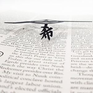 Black metal bookmark resting across an open book, showing a Zen kanji symbol beneath a thin geometric metal design.