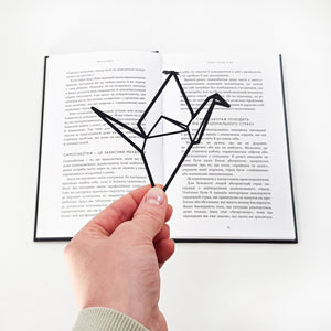 Black metal origami bird bookmark held in hand in front of an open book, showing a minimalist geometric silhouette.