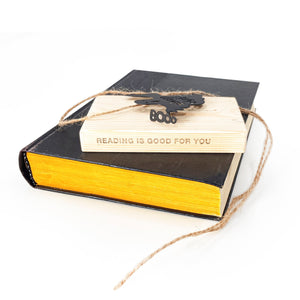 Black steel “Boo” ghost bookmark resting on a wooden block engraved with “Reading is good for you,” placed atop a black book with yellow-edged pages.