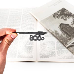 Hand holding a black 'BOO' bookmark over an open book with a Halloween-themed illustration.